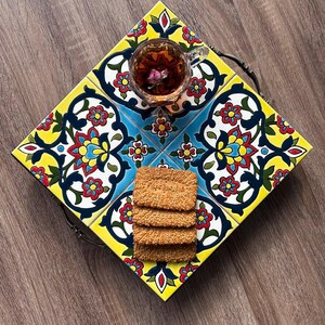 May include: A decorative serving tray with a colorful floral tile design in yellow, blue, red, and white. The tray holds a glass teacup with a flower and a stack of sesame seed crackers. The tray has a dark metal handle.