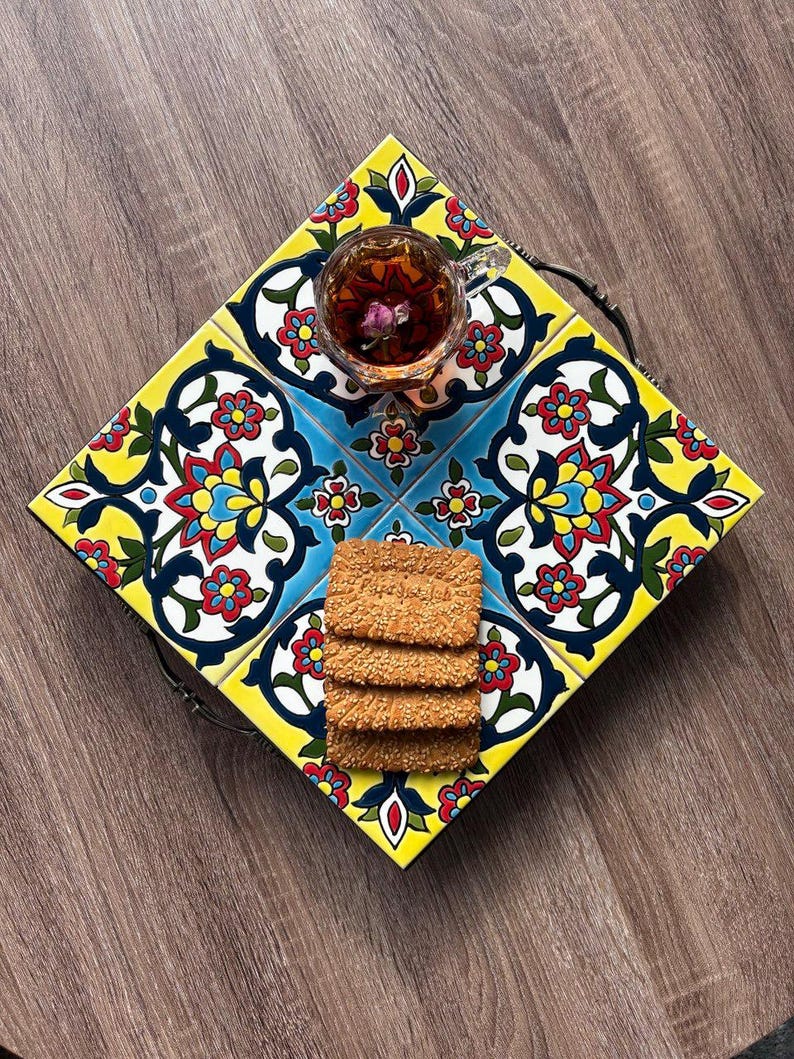 May include: A decorative serving tray with a colorful floral tile design in yellow, blue, red, and white. The tray holds a glass teacup with a flower and a stack of sesame seed crackers. The tray has a dark metal handle.