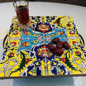 Könnte beinhalten: Ein quadratisches Serviertablett mit vier dekorativen Keramikfliesen in einem lebendigen Blumenmuster in Gelb, Blau, Rot und Grün. Das Tablett hat dunkle Metallgriffe an den Seiten und enthält ein Glas mit dunklem Tee sowie mehrere getrocknete Datteln.