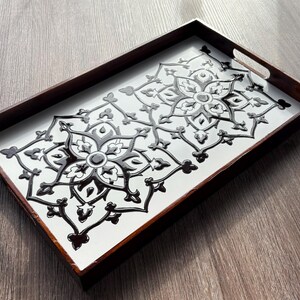 May include: Rectangular wooden serving tray with a white surface and a black floral design. The tray has dark brown edges and integrated handles. The decorative pattern is symmetrical and raised, creating a textured effect.