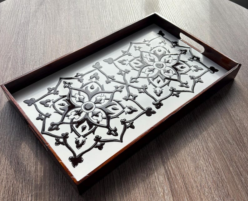 May include: Rectangular wooden serving tray with a white surface and a black floral design. The tray has dark brown edges and integrated handles. The decorative pattern is symmetrical and raised, creating a textured effect.
