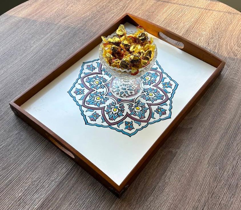 May include: A rectangular wooden serving tray with a white interior featuring a blue, teal, and maroon floral design. A glass bowl filled with wrapped candies sits in the center. The tray has built-in handles.