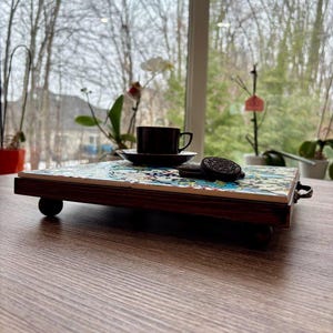 May include: A decorative serving tray with a dark wood frame and a colorful tiled surface. A dark brown teacup and saucer sit on the tray, along with two chocolate sandwich cookies. The tray is on a wooden table.
