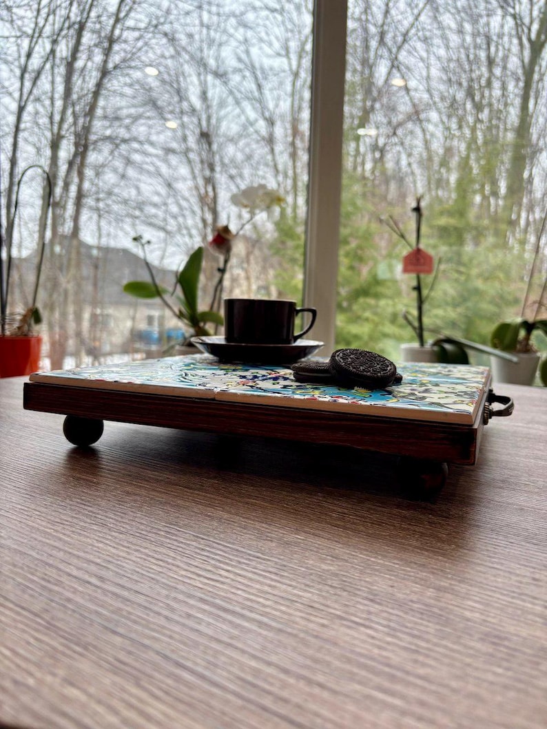 May include: A decorative serving tray with a dark wood frame and a colorful tiled surface. A dark brown teacup and saucer sit on the tray, along with two chocolate sandwich cookies. The tray is on a wooden table.