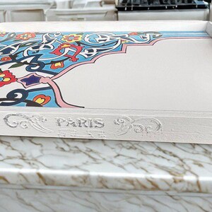 May include: A rectangular white serving tray with handles and decorative corner accents. The tray features a colorful floral and geometric design in blue, red, and yellow. The word "PARIS" is embossed on the side.