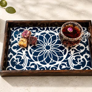 以下が含まれることがあります： 光沢のある白とネイビーブルーの花柄のインレイが施された、ダークウッドの長方形のサービングトレイ。トレイの上には、濃い紅茶が入った透明なガラスのカップと、3つのカラフルな四角いトルコ風の砂糖菓子が置かれています。