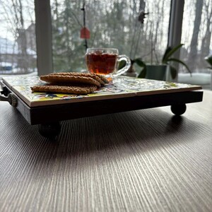 May include: A decorative serving tray with a colorful tile top, dark wooden frame, and small round feet. The tray holds a clear glass teacup filled with tea and a stack of cookies. The background shows a window with a view of trees.