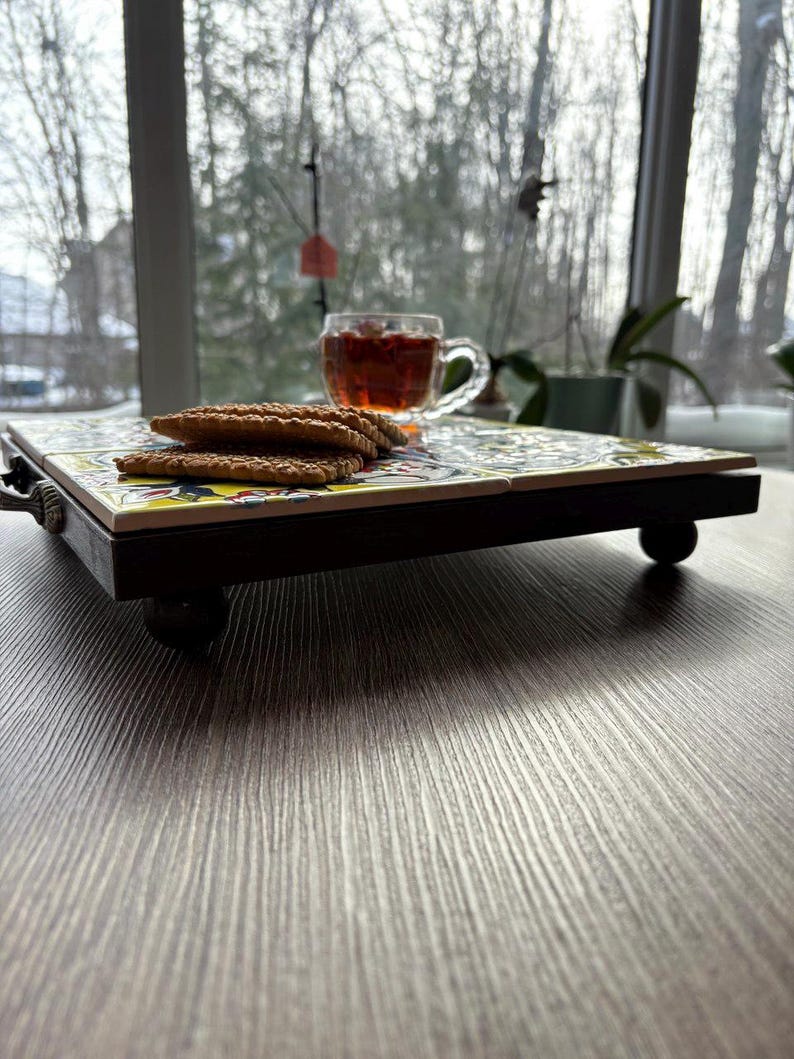 May include: A decorative serving tray with a colorful tile top, dark wooden frame, and small round feet. The tray holds a clear glass teacup filled with tea and a stack of cookies. The background shows a window with a view of trees.