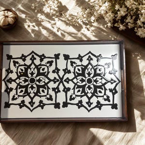 May include: A rectangular wooden serving tray with a white surface featuring a symmetrical black floral design. The tray has a dark brown frame and handles. Decorative elements, including small pumpkins and flowers, are in the background.