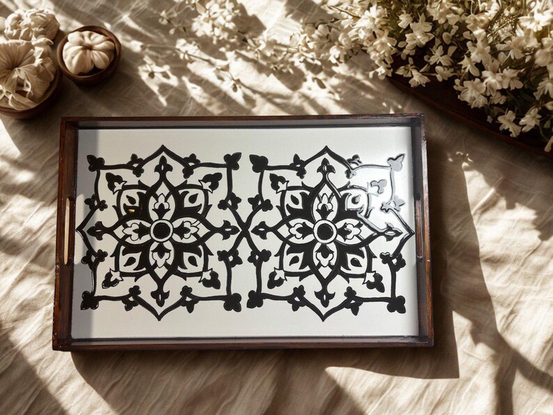 May include: A rectangular wooden serving tray with a white surface featuring a symmetrical black floral design. The tray has a dark brown frame and handles. Decorative elements, including small pumpkins and flowers, are in the background.
