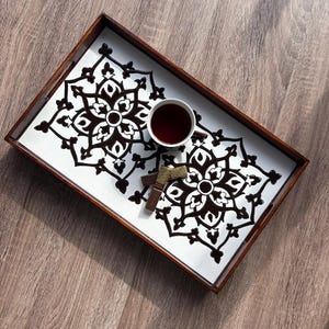 May include: A rectangular wooden serving tray with a white interior featuring a black floral design. A white teacup filled with tea and two pieces of chocolate are on the tray, which sits on a wooden surface.