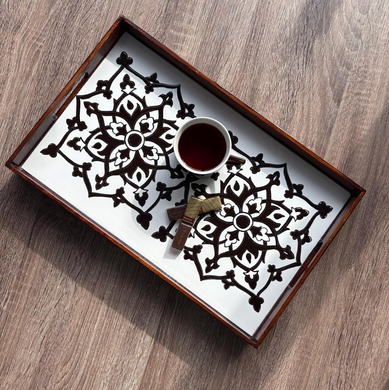 May include: A rectangular wooden serving tray with a white interior featuring a black floral design. A white teacup filled with tea and two pieces of chocolate are on the tray, which sits on a wooden surface.
