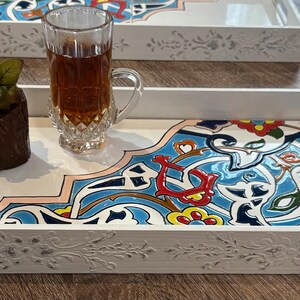May include: White wooden serving tray with a decorative floral design and a colorful, patterned tile insert. The tray holds a glass of tea and a small flower arrangement. Additional trays are visible.