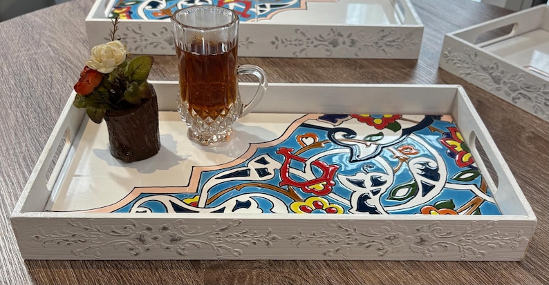 May include: White wooden serving tray with a decorative floral design and a colorful, patterned tile insert. The tray holds a glass of tea and a small flower arrangement. Additional trays are visible.