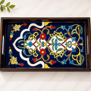 May include: A rectangular wooden serving tray with a dark brown finish and integrated side handles. The base features a vibrant, glossy ceramic tile inlay with a deep blue background and a symmetrical floral pattern in red, yellow, white, and green.