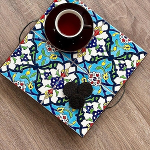 May include: A decorative serving tray with a colorful floral tile design in blue, white, yellow, and red. The tray holds a dark brown teacup and saucer, and three chocolate sandwich cookies. The tray has black metal handles.