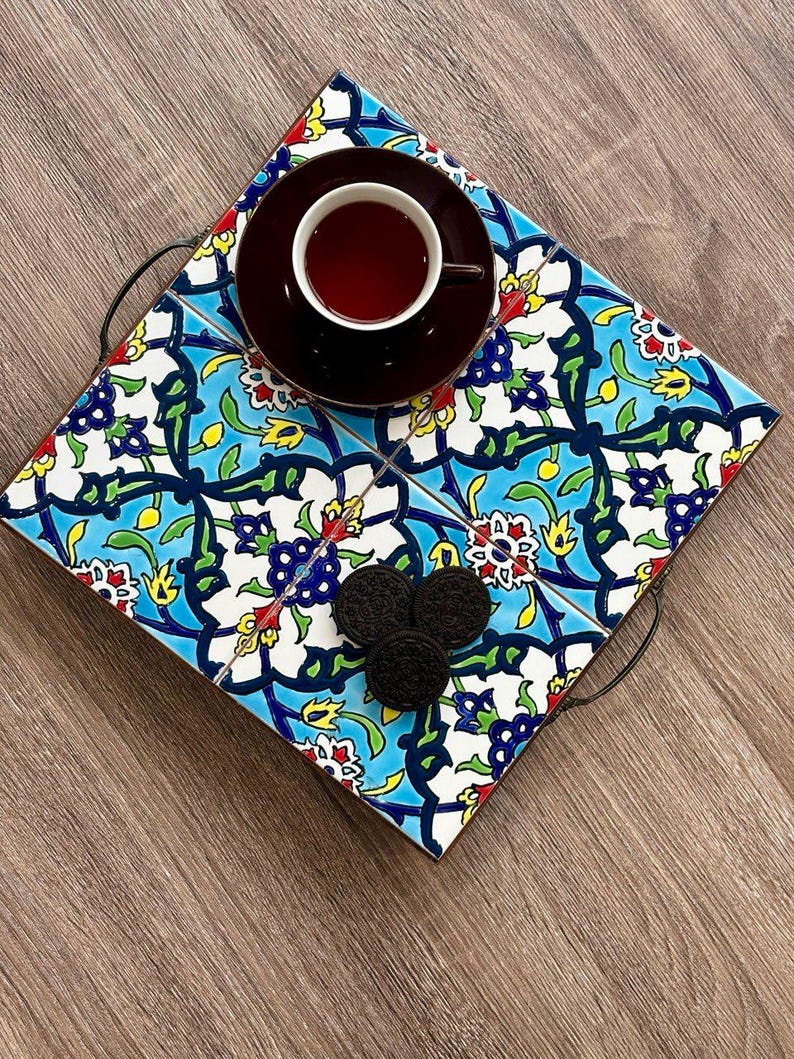 May include: A decorative serving tray with a colorful floral tile design in blue, white, yellow, and red. The tray holds a dark brown teacup and saucer, and three chocolate sandwich cookies. The tray has black metal handles.