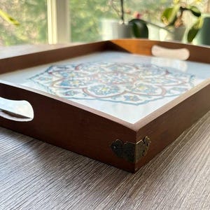 May include: A rectangular wooden serving tray with a decorative floral design. The tray has a dark brown frame with cut-out handles and brass corner accents. The floral design features blue, white, and red colors.