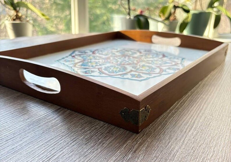 May include: A rectangular wooden serving tray with a decorative floral design. The tray has a dark brown frame with cut-out handles and brass corner accents. The floral design features blue, white, and red colors.