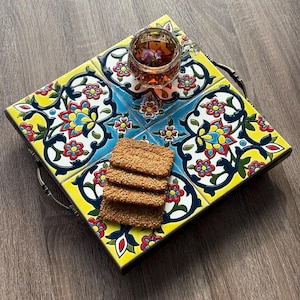 May include: A square serving tray with a colorful floral tile design. The tray has a dark wooden frame and metal handles. A glass of tea with a flower and a stack of crackers are on the tray.