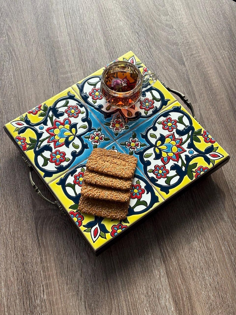 May include: A square serving tray with a colorful floral tile design. The tray has a dark wooden frame and metal handles. A glass of tea with a flower and a stack of crackers are on the tray.
