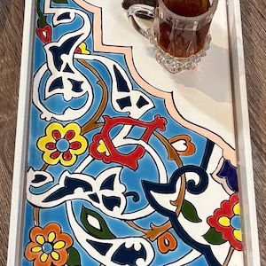 Puede incluir: Bandeja rectangular con marco de madera blanco. La bandeja presenta un diseño de azulejos estampado vibrante en azul, blanco, rojo, amarillo y verde. Un vaso de té y una pequeña planta están en la bandeja.
