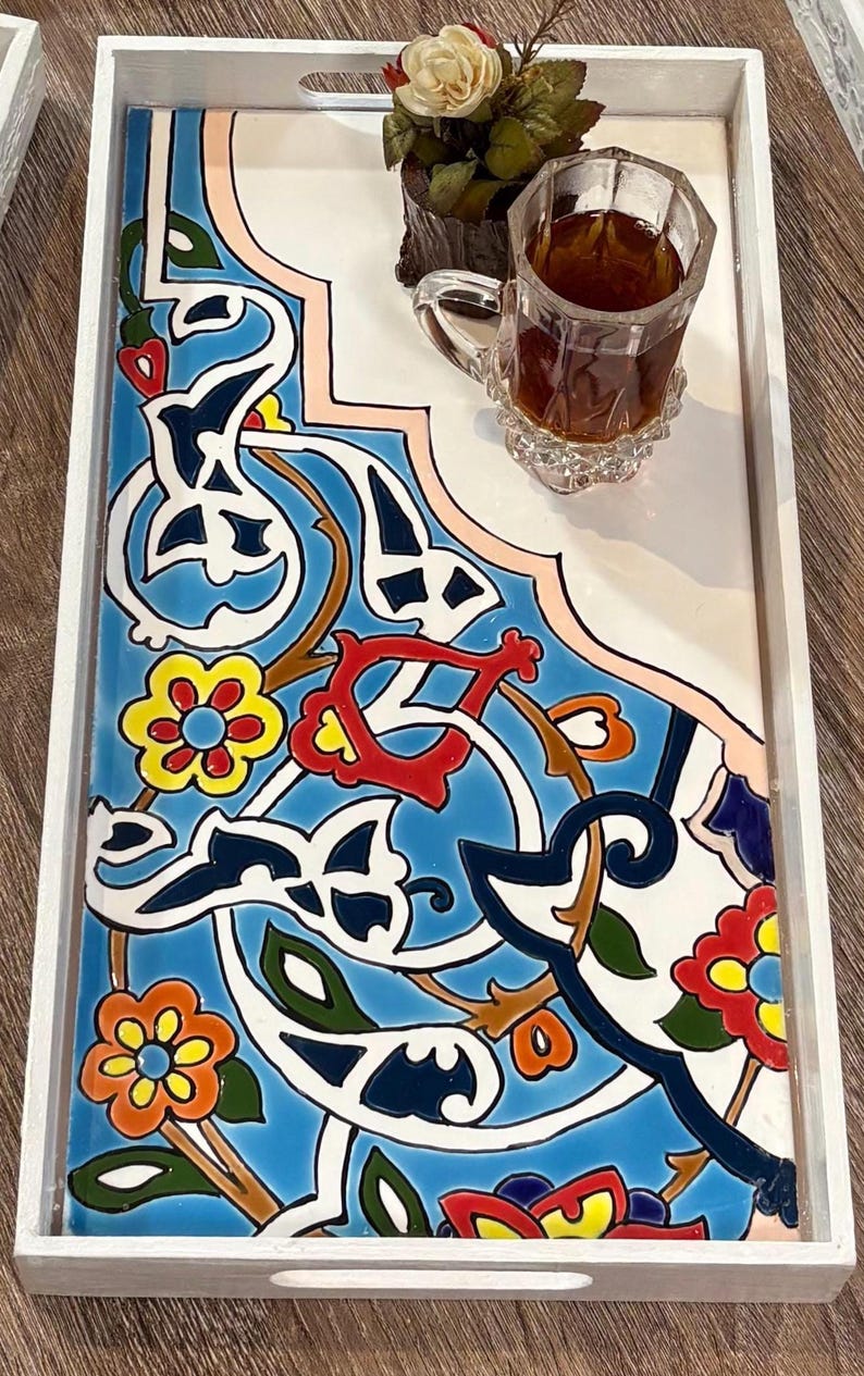 May include: A rectangular serving tray with a white wooden frame. The tray features a vibrant, patterned tile design in blue, white, red, yellow, and green. A glass of tea and a small plant sit on the tray.