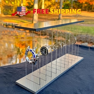 May include: A watch display stand with a black top and a silver base. The stand holds watch components, including a blue and red bezel, a black watch face, and internal mechanisms. The background is a blurred outdoor scene.