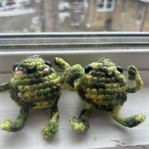 May include: Two handmade crochet frog keychains. The frogs are green and yellow with black eyes and pink cheeks. They have small arms and legs, and are posed in a playful manner. The keychains are hanging on a window sill.