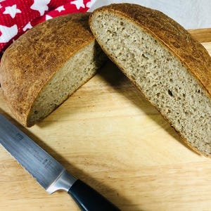 May include: A round loaf of sourdough bread cut into two halves, revealing a light brown, porous crumb and a darker, crusty exterior. A serrated bread knife with a black handle lies diagonally across the wooden cutting board surface.