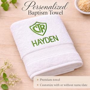 May include: A white cotton towel featuring green embroidery of a shield containing the letters CTR above the name HAYDEN. The text Personalized Baptism Towel is visible at the top, with additional text at the bottom regarding customization.