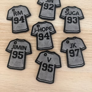Könnte beinhalten: Sieben graue, trikotförmige Aufnäher mit schwarzen Stickrändern und Text. Jeder Aufnäher zeigt einen Namen und eine zweistellige Zahl: RM 94, JIN 92, SUGA 93, J-HOPE 94, JIMIN 95, V 95 und JK 97. Jeder Aufnäher hat oben eine kleine kreisförmige Öse.