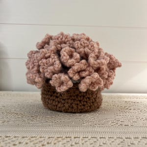 May include: A handmade crochet flower arrangement in a brown pot. The flowers are a soft pink color, and the pot is a darker brown. The arrangement is sitting on a cream-colored lace tablecloth.