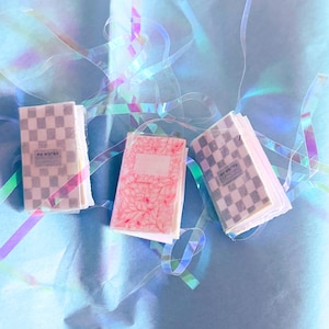 Puede incluir: Tres cuadernos pequeños con cubiertas decorativas. Dos cuadernos tienen un patrón de cuadros grises y blancos, y uno tiene un diseño floral rosa. Los cuadernos están sobre una superficie azul con cintas iridiscentes.