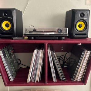 May include: A home entertainment setup featuring a red wooden shelf with a turntable, black speakers with yellow cones, and vinyl records. The shelf is mounted on a wall, creating a space for music and audio equipment.