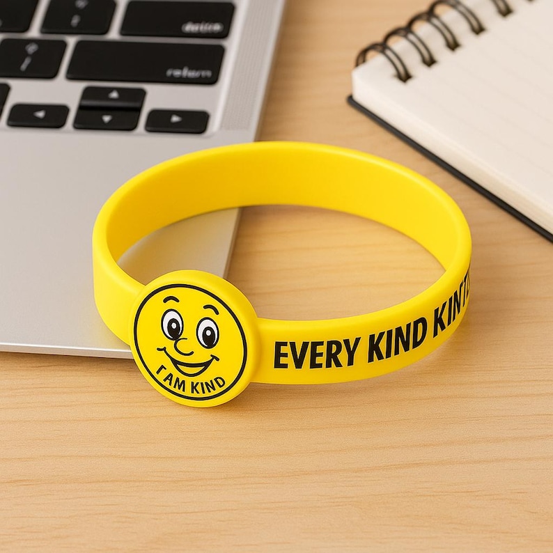Puede incluir: Una pulsera de silicona amarilla con el texto "EVERY KIND KIND" y una carita sonriente con el texto "I AM KIND". La pulsera est&aacute; sobre una superficie de madera cerca de un port&aacute;til y un cuaderno.