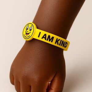 Puede incluir: Una pulsera de goma amarilla con las palabras "I AM KIND" en letras negras. La pulsera presenta una cara sonriente redonda con la palabra "DAMIKIND" en el lateral. La pulsera est&aacute; en la mu&ntilde;eca de un ni&ntilde;o.
