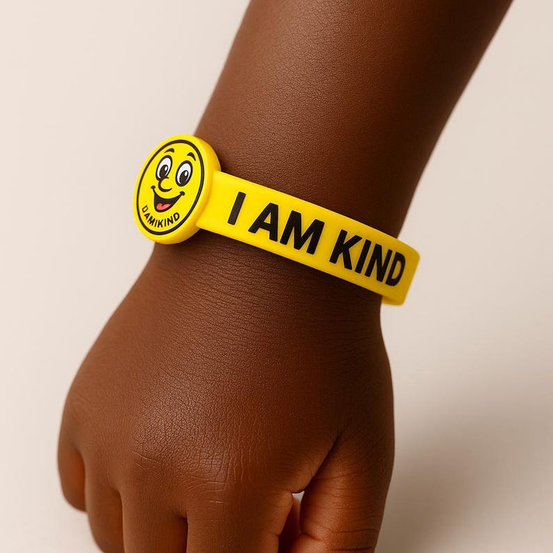 Puede incluir: Una pulsera de goma amarilla con las palabras "I AM KIND" en letras negras. La pulsera presenta una cara sonriente redonda con la palabra "DAMIKIND" en el lateral. La pulsera est&aacute; en la mu&ntilde;eca de un ni&ntilde;o.