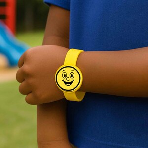 Puede incluir: Una pulsera de silicona amarilla con un dise&ntilde;o de cara sonriente. La esfera del reloj es un c&iacute;rculo con un contorno negro, con ojos, nariz y boca sonriente. La pulsera est&aacute; sujeta a una mu&ntilde;eca.