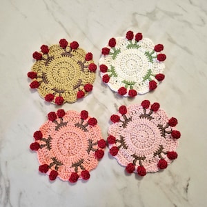 Puede incluir: Cuatro posavasos de ganchillo hechos a mano en tonos rosa, crema y beige, cada uno con un centro en espiral y un borde festoneado. Los bordes están adornados con pequeños capullos de flores rojos de ganchillo. Un artículo decorativo y funcional.