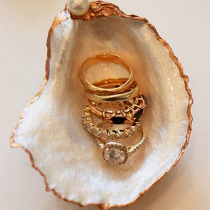 Puede incluir: Un conjunto de anillos de oro dentro de una concha de ostra decorativa. La concha tiene un interior nacarado y un borde dorado. Una sola perla se encuentra en el borde de la concha. Los anillos varían en diseño, incluyendo uno con diamantes.