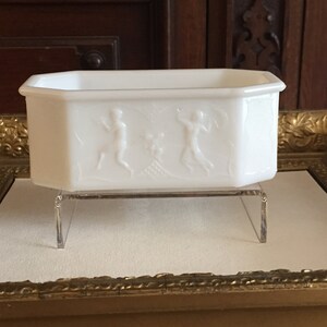 May include: White octagonal glass dish with an embossed design of three figures. The dish is on a clear acrylic stand.