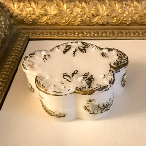 May include: A white porcelain trinket box with a gold and white floral design. The box has a scalloped edge and a lid with a raised design.