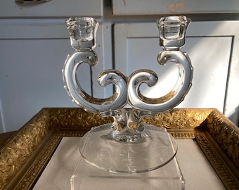 May include: A clear glass candle holder with two candle cups and a swirling design. The holder is on a white surface.