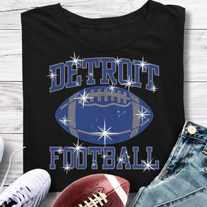 May include: Black t-shirt with the words "DETROIT FOOTBALL" in a sparkling, rhinestone-like font. A blue football graphic is centered on the shirt, also with a rhinestone design. A brown football, white sneakers, and blue jeans are in the image.