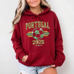 May include: Red hoodie featuring the customized Portugal rhinestone soccer graphic. The high-shine stones bring a trendy, athletic vibe to the classic hooded garment with a front pouch pocket.