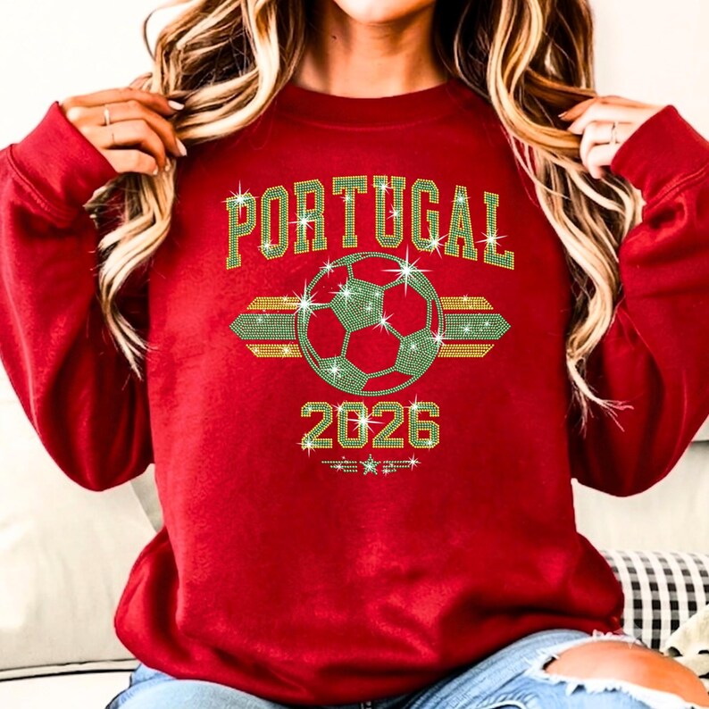 May include: Red crewneck sweatshirt showcasing a brilliant 'Portugal 2026' rhinestone football arrangement. The shimmering yellow and green glass stones offer a premium, high-contrast sporty look on a cozy heavy-blend fabric.