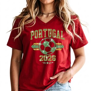 May include: Red V-neck t-shirt highlighting the arched 'PORTUGAL' script and central rhinestone soccer ball. The elegant neckline beautifully frames the sparkling two-tone layout on soft Bella+Canvas fabric.