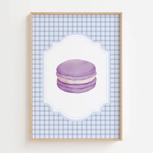 May include: A framed print featuring a watercolor illustration of a purple macaron with white filling. The macaron is centered on a white, ornate label against a light blue gingham pattern background. The frame is a light wood color.