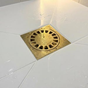 Könnte beinhalten: Ein quadratischer, goldfarbener Duschablauf mit kreisförmigem, geschlitztem Design. Der Ablauf ist in weiße Fliesen verlegt. Der Ablauf hat eine strukturierte Oberfläche und ist von Wassertropfen umgeben.